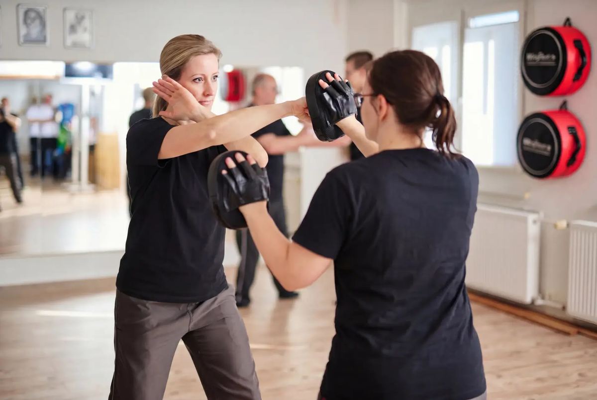 Zwei Frauen üben in einer Kampfsportschule Schläge auf Pratzen, im Hintergrund weitere Trainierende